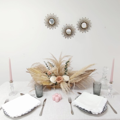 Mesa de jantar posta com arranjo floral seco, velas rosas, pratos brancos, talheres e copos cinzentos, e três espelhos dourados na parede