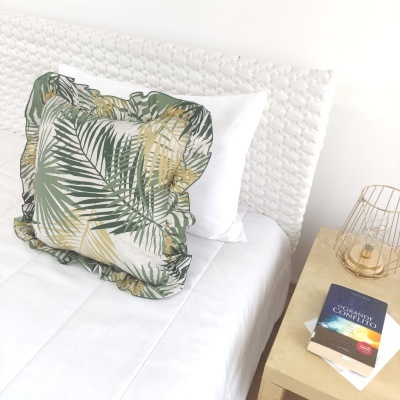 almofada quadrada com padrão de folhas verdes e amarelas numa cama branca ao lado de mesa de cabeceira com candeeiro dourado e livro