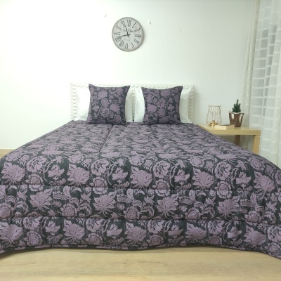 Conjunto de cama com colcha e almofadas florais roxas e cinza escuro num quarto branco.