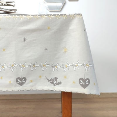 Toalha de mesa clara com padrão de estrelas douradas e cinzentas, corações cinzentos com flores e aves em borda, sobre mesa de madeira clara