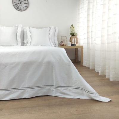 Colcha branca com bordado cinza numa cama em quarto claro com cortinas e mesa de cabeceira