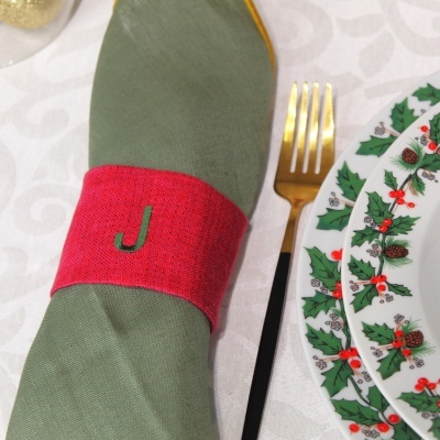 Mesa posta com guardanapo verde e argola vermelha com letra J, garfo dourado e prato com padrão natalino