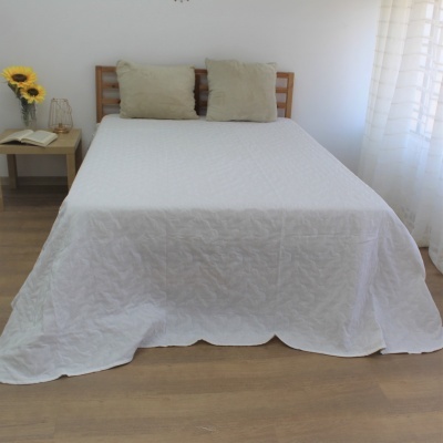 Cama de casal com colcha branca e almofadas bege num quarto minimalista com criado-mudo e girassóis.