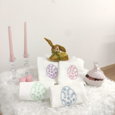Toalhas de banho brancas com bordados coloridos de ovo da Páscoa, coelho decorativo, velas cor-de-rosa em castiçais transparentes e caixa decorativa de coelho sobre superfície felpuda branca.