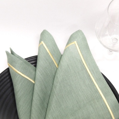 Guardanapo de tecido verde com borda dourada sobre prato preto e copo transparente