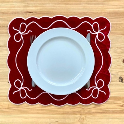 Prato branco redondo sobre individual vermelho de veludo com laços brancos numa mesa de madeira clara