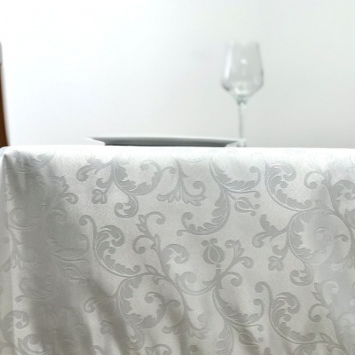 Toalha branca com padrão floral em mesa com cadeira de madeira, prato e copo ao fundo