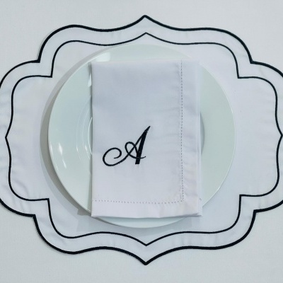 Jogo americano branco com bordado preto e guardanapo branco bordado com a letra A sobre prato branco
