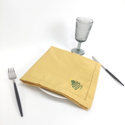 Guardanapo amarelo com bordado verde sobre prato branco, com talheres pretos e copo de vidro cinza