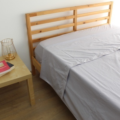 Cama de madeira clara com roupa de cama cinzenta e mesa de cabeceira com livros e lanterna dourada