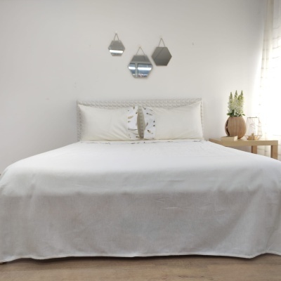 Cama com roupa de cama branca e almofadas decoradas, espelhos geométricos na parede e mesa lateral com vaso de plantas.