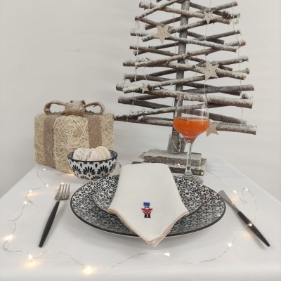 Mesa de Natal com prato preto e branco, guardanapo branco bordado, talheres pretos, copo com bebida alaranjada, árvore de Natal em ramos secos e presente decorativo.