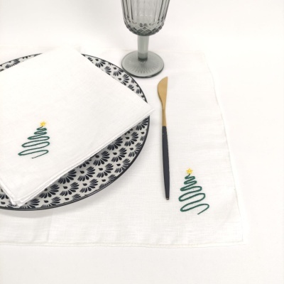Guardanapo e toalha brancos com bordado de árvore de Natal verde e estrela amarela, prato decorado, faca preta e dourada, copo cinzento.