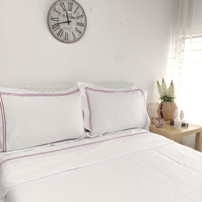 Cama com roupa branca e linhas vermelhas, relógio cinza na parede e mesa de cabeceira com vasos