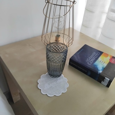 Frasco de vidro cinzento com padrão hexagonal sobre descanso de copo branco, livro e lâmpada de metal numa mesa de madeira clara