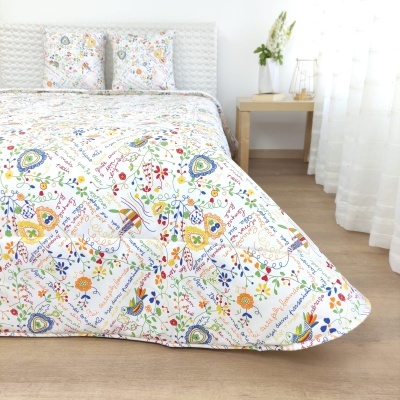 Cama com edredão e almofadas florais coloridas com texto, mesa de cabeceira e cortinas brancas.