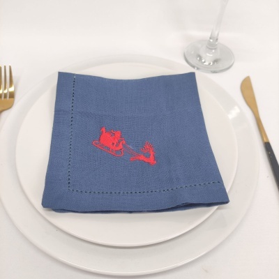 Guardanapo azul com bordado vermelho, em pratos brancos com talheres dourados e copo de vidro