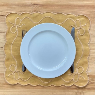 Prato branco sobre jogo americano amarelo com bordado de laços, garfo e faca ao lado, sobre mesa de madeira clara