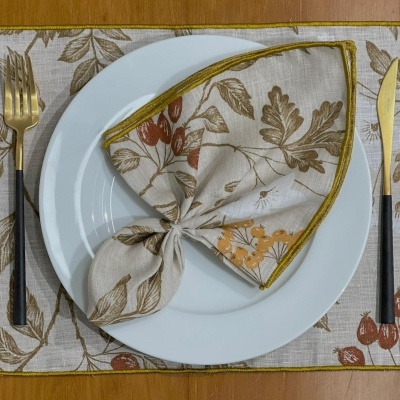 Jogo americano e guardanapo com padrão de folhas e frutos, prato branco, talheres preto e dourado sobre mesa de madeira