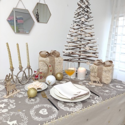 Mesa decorada para Natal com toalha cinza estampada, pratos brancos, castiçal dourado, presentes dourados, bola dourada e árvore de ramos brancos