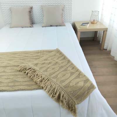 Cobertor bege com padrão trançado e franjas sobre cama branca com almofadas e mesa de madeira clara ao lado