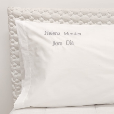 Fronha branca bordada com nome e mensagem sobre cama com cabeceira acolchoada branca