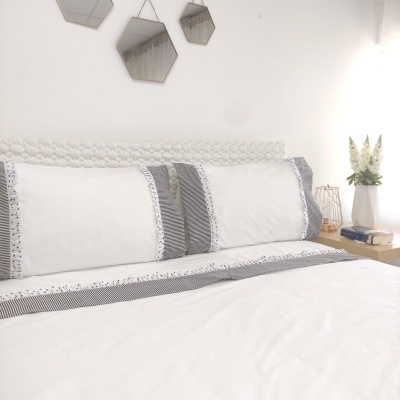 Conjunto de roupa de cama branco com detalhes cinzentos e bordados, cabeceira tecida, espelhos hexagonais e mesa de cabeceira com flores