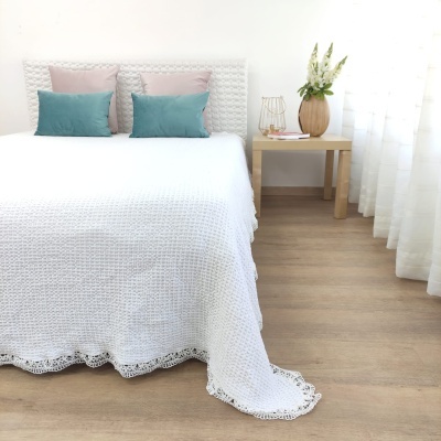 Cama com colcha branca e almofadas coloridas, mesa de cabeceira com vaso de flores, ambiente luminoso