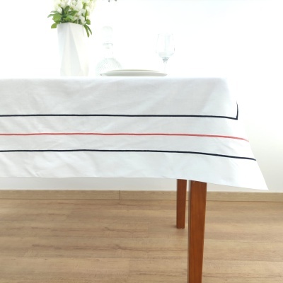 Toalha de mesa branca com riscas pretas e vermelhas sobre mesa de madeira com vaso, prato e copo.