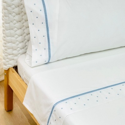 Lençóis e fronhas brancas com bordado azul claro em cama de madeira e cabeceira trançada branca.