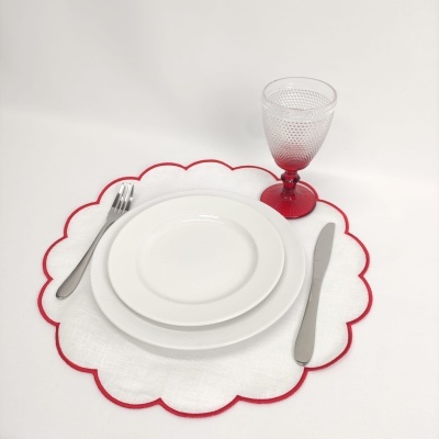 Conjunto de jantar com pratos brancos, talheres metálicos, individual branco com borda vermelha, e taça com base vermelha.