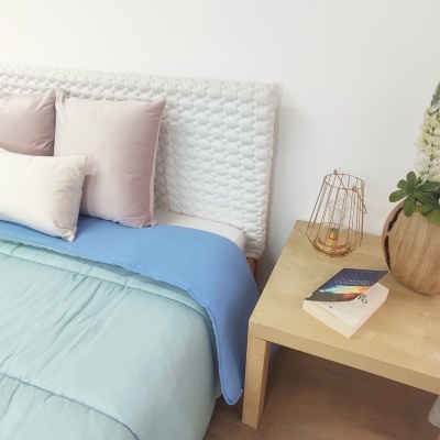 Cama com cabeceira branca trançada, manta azul, almofadas rosa e creme, mesa de madeira com vaso, vela e livros.