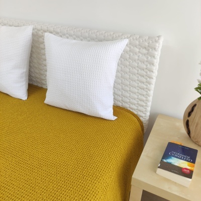Cama com cabeceira branca, almofadas brancas, cobertor amarelo e mesa de cabeceira com vaso e livro
