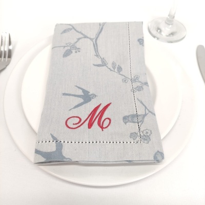 Guardanapo cinzento com padrão de aves e flores e letra 'M' bordada em vermelho