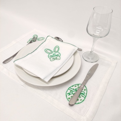 Jogo de jantar com toalhete e base de tecido com bordados verdes de coelho e ovo de páscoa, prato branco, talheres metálicos e copo de vidro
