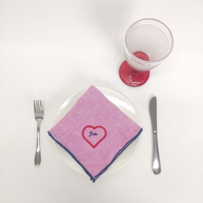 Jogo de mesa com guardanapo rosa com borda azul e nome 'João', prato branco, talheres metálicos e copo com pé vermelho