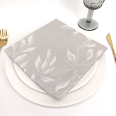Guardanapo cinzento claro com padrão de folhagem em branco sobre pratos brancos com talheres dourados e copo cinzento