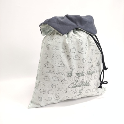Saco de pano cinza claro com padrão de coelhos, cordão preto e parte superior cinza escuro com texto verde.