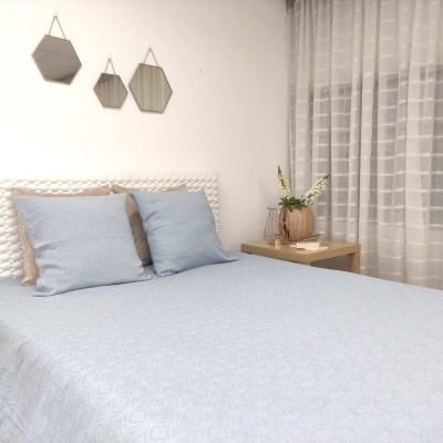 Cama com roupa de cama azul claro e almofadas, cabeceira branca texturizada e mesa de cabeceira com vaso de plantas.