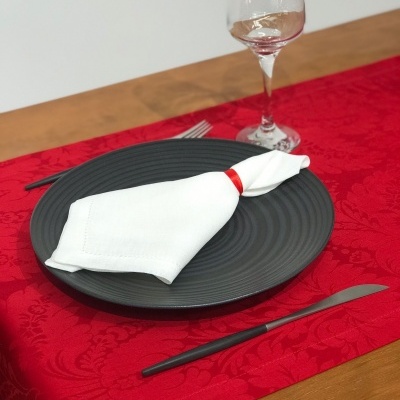 Mesa posta com prato preto, talheres pretos, guardanapo branco com argola vermelha, copo de vinho e toalha de mesa vermelha floral