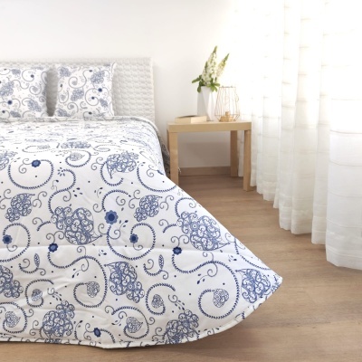 Cobertura de cama com padrão azul e branco floral, mesa de madeira com vaso de flores e cortinas brancas translúcidas