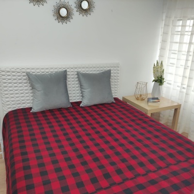 Cama com colcha xadrez vermelha e preta, almofadas cinza, cabeceira branca acolchoada e mesa de madeira com decoração.