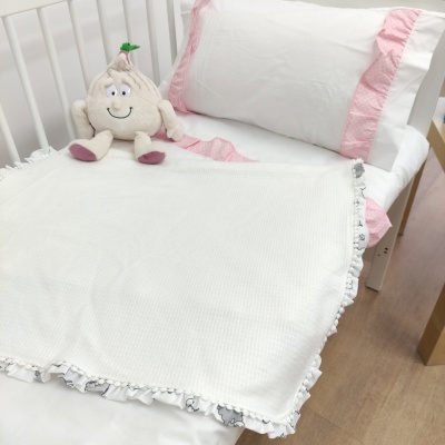Cobertor branco de malha com frisos estampados cinzentos na borda, cama branca de grades com almofada branca com fita rosa e peluche de alho sorridente