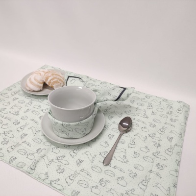 Conjunto de chávena, pires, prato com pastéis, colher e pano de mesa com desenhos de animais.