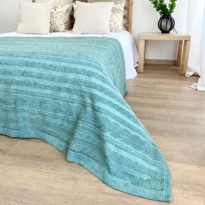 Colcha azul tricotada sobre cama com almofadas e mesa lateral de madeira clara