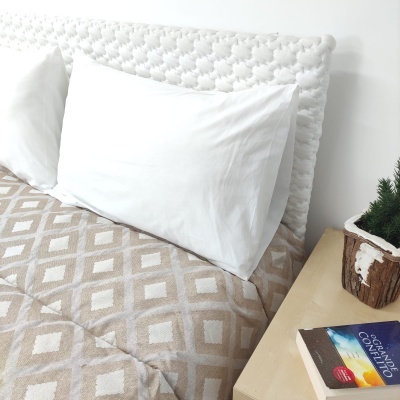 Cama com cabeceira branca texturizada, almofadas brancas e edredão bege com padrão geométrico ao lado de mesinha com vaso e livro.