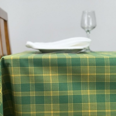Mesa com toalha xadrez verde e amarela com pratos brancos, cadeira de madeira e copo de vidro ao fundo