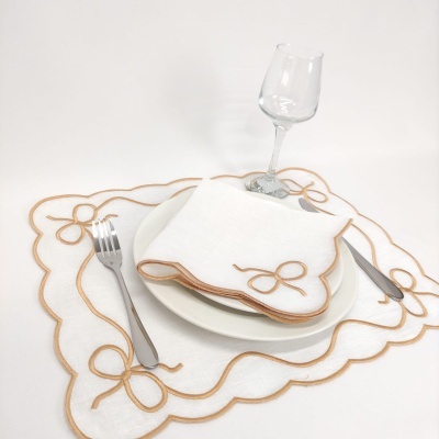 Guardanapo e passatempo em tecido branco com bordado castanho em forma de laço, utensílios metálicos, prato branco e copo de vidro.