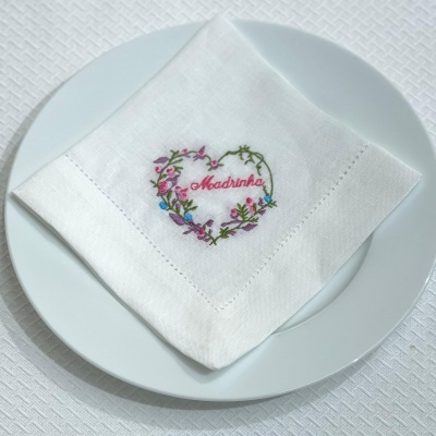 Guardanapo branco bordado com a palavra 'Madrinha' em coração floral sobre prato branco