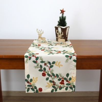 Caminho de mesa natalício com ramos verdes, bagas vermelhas, e sinos dourados, com figuras de veados e uma árvore de Natal pequena em leiteira decorativa.